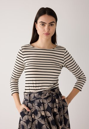 Frau mit langem dunklem Haar trägt ein beige-schwarzes gestreiftes Langarmshirt und einen hoch taillierten Rock mit kreisförmigen Fächer-Mustern, Hände in den Taschen.