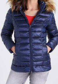 Doudoune bleu marine avec une fermeture éclair frontale, un motif matelassé horizontal et une capuche amovible en fausse fourrure. Comprend deux poches avant.