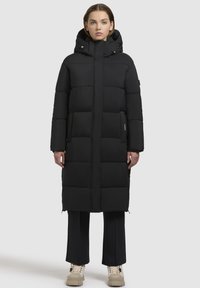 Sort polstret lang frakke med hætte. Lodret quiltet design, to sidelommer og lynlås lukning. Båret over sorte bukser og beige sko.