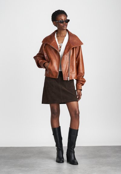 Bruine leren kort jack met brede kraag, ritssluiting en zijzakken. Gecombineerd met een witte blouse en een bruine suède rok. Zwarte laarzen tot op de knie.