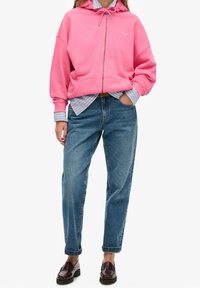 Vrouw in een roze ritsjack over een gestreept blauwe shirt, blauwe spijkerbroek en donkerbruine instappers, staand met één hand in de zak.