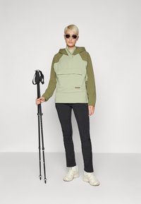 Lys grønn og olivengrønn to-tonet jakke med hette og frontlomme, parret med svarte bukser og beige joggesko, holder turstaver.