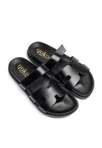 Sandales noires avec une texture lisse, dotées de larges lanières, d'un design découpé et de détails de logo doré. Semelle plate, style moderne.