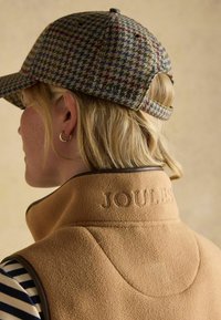 Beige Fleece-Weste mit hohem Kragen, mit eingeprägtem "JOULES"-Logo. Graue, karierte Mütze mit mehrfarbigen Akzenten, rückwärts getragen. Gestreifter Stoff darunter.