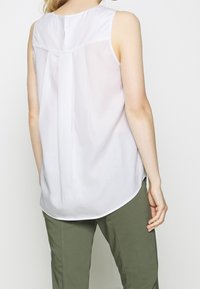 Mujer vestida con blusa sin mangas blanca y pantalones verdes oliva, mostrada de espaldas contra un fondo blanco liso.