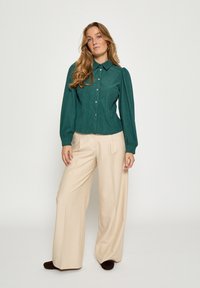 Žalvarinės spalvos marškiniai su apykakle ir pūstomis rankovėmis, derinami su aukšto liemens kreminės spalvos plačiomis kelnėmis ir tamsiais batais.