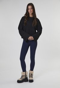 Suéter negro con puños de canalé anchos, leggings azul marino y botas negras con forro de pelo y cordones. El modelo está de pie contra un fondo neutro.