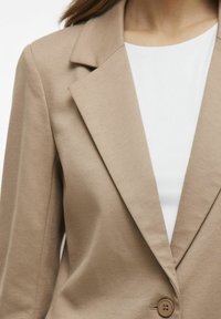 Blazer beige con rever a punta, chiusura a un bottone e tessuto texturizzato. Indossato sopra una maglietta bianca, che mette in risalto il design sartoriale.