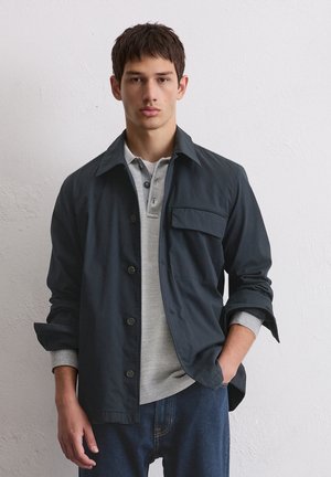 Dark navy jacket with a collar, button front, and one chest pocket over a light gray polo. Model wears denim jeans. Smooth fabric texture.