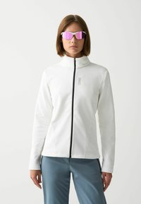 Giacca bianca con zip e colletto alto, texture liscia e zip nera. Il modello indossa occhiali da sole rosa specchiati e pantaloni azzurri chiari.