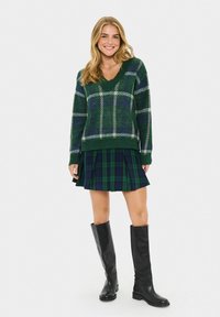 Lächelnde Frau trägt einen grün karierten Pullover, einen gefältelten Schottenrock und schwarze kniehohe Stiefel, steht vor einem schlichten weißen Hintergrund.
