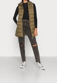 Olivgrüne gesteppte Weste mit Kapuze über einem schwarzen, gerippten Oberteil, kombiniert mit dunklen, zerrissenen Jeans und schwarz-weiß gemusterten High-Top-Sneakers.