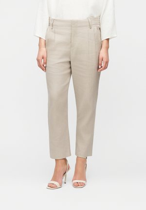 Kvinde iført beige skræddersyede bukser, hvid langærmet top og beige højhælede sandaler med åben tå, stående mod en ensfarvet baggrund.