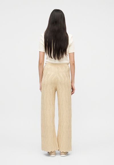 Γυναίκα με μακριά σκούρα μαλλιά στέκεται με την πλάτη γυρισμένη, φορώντας λευκό κοντομάνικο τοπ, μπεζ υφασμάτινο παντελόνι με φαρδιά πόδια και αθλητικά παπούτσια.