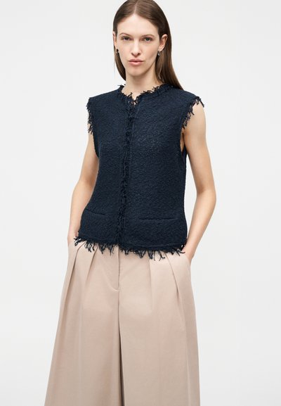 Γυναίκα που φοράει αμάνικο μπλε σκούρο υφασμάτινο γιλέκο με ξεφτισμένες άκρες και μπεζ παντελόνι με φαρδιά μπατζάκια και ψηλή μέση, στέκεται με τα χέρια στις τσέπες.