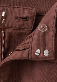 Tela de mezclilla marrón con cierre de cremallera, botón y detalles de hardware metálico. Superficie texturizada con costuras visibles.