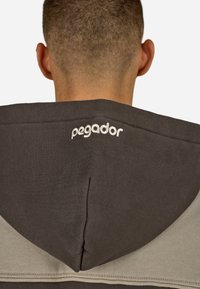 Hætte sweatshirt i grå og beige stof. Har et broderet logo "pegador" på hætten og en glat tekstur.