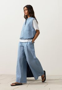 Femme marchant avec un gilet sans manches rayé bleu et blanc et un pantalon à jambes larges, avec une chemise blanche à manches courtes et des tongs noires sur un sol carrelé.