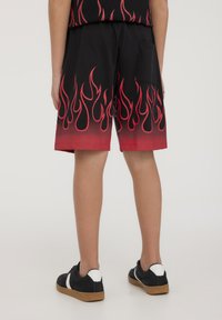 Pantaloni corti neri con motivi di fiamme rosse all'orlo, dotati di una tasca posteriore e un fit rilassato, abbinati a scarpe nere con dettagli bianchi.