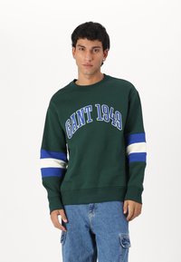 Groene sweatshirt met het "GANT 1949" logo in blauw, witte en blauw gestreepte accenten op de mouwen. Zachte textuur, klassiek model met ronde hals.