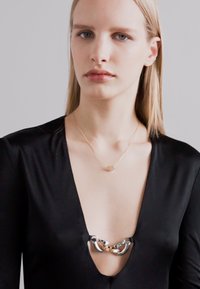 Črna obleka z globokim V-izrezom, ki ima metalno verižico kot okras na izrezu ter nežno zlatno verižico z majhnim obeskom.