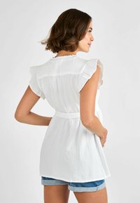 Biele, ľahké rúcho s volánikmi na rukávoch, zaväzovaním v páse a textúrovanou látkou. Pohľad zozadu ukazuje voľný strih a jemné splývanie.