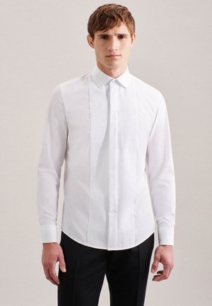 Jeune homme portant une chemise blanche à manches longues avec un devant plissé et un pantalon noir, debout devant un fond clair uni.