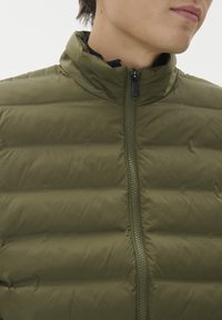 Pogled od blizu na osobu koja nosi jaknu maslinaste boje s punjenjem, frontnim patentnim zatvaračem i visokim ovratnikom, glavom lagano okrenutom na stranu.