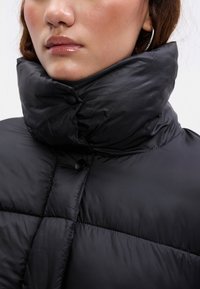 Casaco acolchoado preto com colarinho alto, apresentando fechos de botão e textura almofadada, projetado para proporcionar calor e conforto.