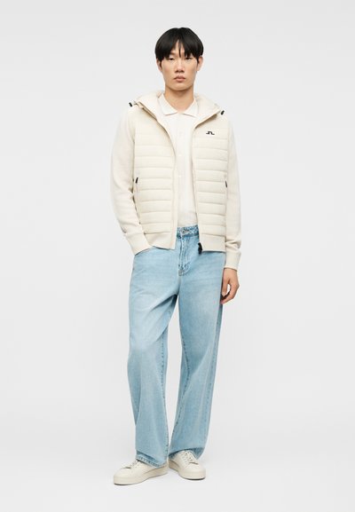 Νέος άνδρας στέκεται φορώντας εκρού καπιτονέ γιλέκο με κουκούλα, εκρού μακρυμάνικο πουκάμισο, ανοιχτό μπλε τζιν με φαρδιά πόδια και εκρού αθλητικά παπούτσια, πάνω σε απλό φόντο.