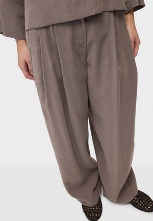 Pantaloni color taupe a vestibilità ampia con pieghe frontali, abbinati a un top coordinato e scarpe basse nere traforate, indossati da una persona in piedi.