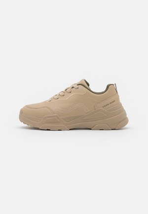 Beżowe sneakersy na sznurówki z teksturowaną syntetyczną cholewką, kontrastującymi zielonymi akcentami i grubą, lekką gumową podeszwą.