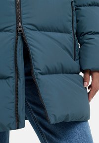 Giubbino imbottito in teal con finitura lucida, dotato di zip full-length e tasche laterali, abbinato a jeans in denim.