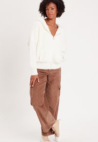 Weißer Kapuzenpullover mit einem flauschigen Textur und Fronttaschen, kombiniert mit braunen Corduroy-Cargo-Hosen und beigen Sneakers.