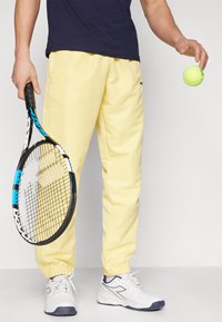 Lys gule joggebukser med elastiske mansjetter, kombinert med en marineblå t-skjorte. Personen holder et tennisracket og en lys gul tennisball.