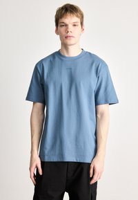 Μπλε βαμβακερό φανελάκι με λαιμόκοψη, κοντά μανίκια και μια διακριτική λογότυπη κεντημένη στο στήθος. Απλή, χαλαρή γραμμή.