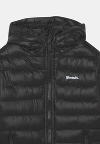 Svart puffjacka med huva, med dragkedjestängning och kviltat mönster. Logotyp "Bench." i vitt på vänster bröst. Slät textur.