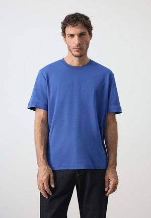 Homme portant un t-shirt bleu uni à manches courtes et un pantalon foncé, debout devant un fond clair uni.