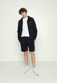 Jonge man staat in een zwart zip-up hoodie, zwarte shorts, wit t-shirt, witte sokken en witte sneakers tegen een effen lichte achtergrond.