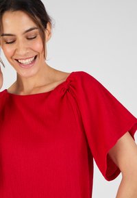 Blusa rossa con texture, dotata di maniche corte e ampie e un dettaglio alla scollatura arricciato. Il tessuto presenta un leggero motivo a righe.
