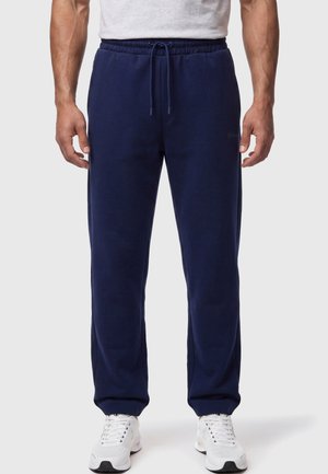 Mann trägt marineblaue Jogginghose mit Kordelzug in der Taille und weiße Sneakers, steht vor schlichtem hellem Hintergrund.