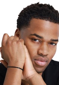 Bracciale a catena nera poggia su un polso, caratterizzato da un design elegante e minimalista con maglie intrecciate. Finitura metallica liscia con una sottile lucentezza.