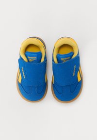 Zapatillas de niño Reebok en azul y amarillo con una correa de velcro, fabricadas en un tejido suave, con suela de goma y detalles de costura en contraste.