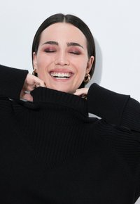 Maglione a collo alto nero a coste con piega prominente, caratterizzato da dettagli testurizzati e maniche lunghe, indossato da una persona sorridente con le mani appoggiate sul collo.