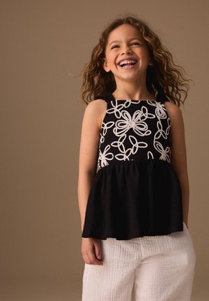 Chica con cabello rizado sonriendo ampliamente, lleva una camiseta sin mangas negra con bordado floral blanco y pantalones blancos sobre un fondo marrón liso.