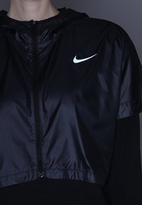 Veste courte noire à capuche en nylon brillant, avec une fermeture éclair frontale et un petit logo Nike blanc sur la poitrine gauche.