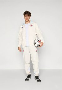 Αθλητικό σακάκι και παντελόνια σε ανοιχτό μπεζ χρώμα, και τα δύο με λογότυπα. Το σακάκι έχει μπλε εσωτερική επένδυση· το άτομο κρατά μια ασπρόμαυρη μπάλα ποδοσφαίρου.