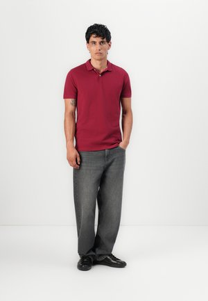 Homme adossé à un mur blanc, portant un polo bordeaux, un jean gris ample et des baskets noires, avec une main dans la poche.