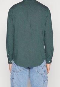 Persoon gekleed in een donkergroen shirt met lange mouwen en lichte spijkerbroek, weergegeven van de achterkant tegen een effen achtergrond.