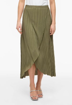 Femme portant une jupe portefeuille plissée vert olive associée à un haut blanc et des sandales argentées à talons hauts avec brides, debout sur un fond blanc.